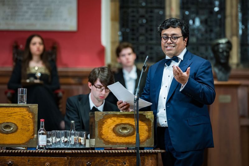 Emad Mostaque speaking at the despatch box in the Oxford Union chamber, closing for the Opposition, with the tellers seated behind him.